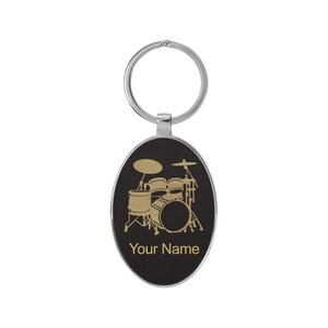 May include: Oval-shaped keychain with a silver-colored ring. The black surface features a gold-colored illustration of a drum set and the text "Your Name". A personalized gift for drummers.