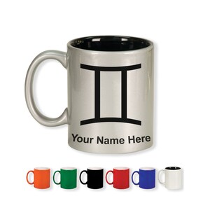 May include: A silver-colored mug with a black interior and handle. The mug features a black Gemini zodiac symbol and the text "Your Name Here". Several colorful mugs are displayed below.