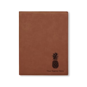 May include: A brown leatherette journal with a black pineapple design and the text "Your Name Here" engraved on the front.