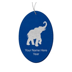 May include: A blue oval-shaped ornament with a silver elephant design. The ornament has the text "Your Name Here" and "Year" in white. A silver string is attached for hanging.