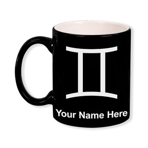 May include: Black ceramic mug with a white interior and handle. The mug features a white Gemini zodiac symbol and the text "Your Name Here" in white. The mug is designed for beverages.