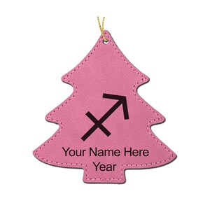May include: A pink Christmas tree ornament with a black Sagittarius symbol and the text "Your Name Here Year". The ornament is made of a textured material and has a gold string for hanging.