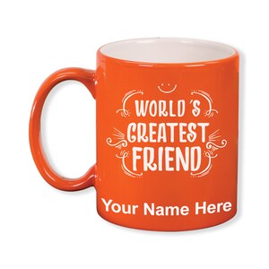 K&ouml;nnte beinhalten: Eine orangefarbene Keramiktasse mit wei&szlig;em Innenraum und Henkel. Die Tasse tr&auml;gt den wei&szlig;en Text "WORLD'S GREATEST FRIEND", mit Platz f&uuml;r Personalisierung darunter. Ein kleines Smiley befindet sich &uuml;ber dem Text.