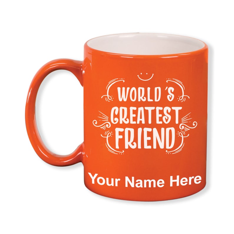 K&ouml;nnte beinhalten: Eine orangefarbene Keramiktasse mit wei&szlig;em Innenraum und Henkel. Die Tasse tr&auml;gt den wei&szlig;en Text "WORLD'S GREATEST FRIEND", mit Platz f&uuml;r Personalisierung darunter. Ein kleines Smiley befindet sich &uuml;ber dem Text.