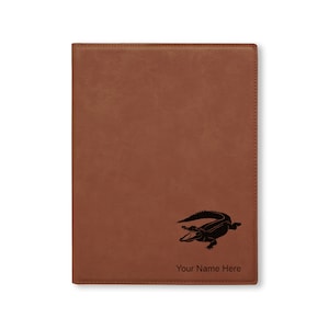 May include: A brown leatherette journal with a black alligator silhouette and the text "Your Name Here" engraved on the front.