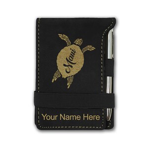 May include: Black leatherette notebook with a gold-colored sea turtle design and the word "Maui" inscribed on the turtle's shell. The notebook has a black elastic band closure and a pen holder. The text "Your Name Here" is engraved on the front of the notebook.