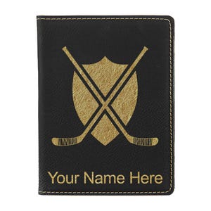 May include: Black passport holder with gold stitching and a gold hockey-themed design. The design features crossed hockey sticks and a shield. The text "Your Name Here" is printed in gold below the design.