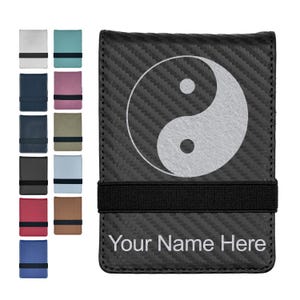 May include: A black carbon fiber textured wallet featuring a white yin-yang symbol on the front and a horizontal black elastic strap. The text "Your Name Here" is printed in white at the bottom. A column of various colored wallet options is shown.