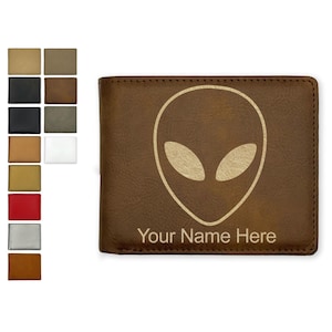 May include: A brown bi-fold wallet featuring a light-colored alien head graphic centered on the front cover. Below the alien graphic, the text "Your Name Here" is printed in a simple, sans-serif font. A color palette of options is shown.