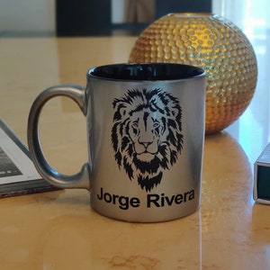 May include: Silver-colored mug with a black interior and a handle. The mug features a black lion's head design and the name "Jorge Rivera" printed below the lion. The mug is sitting on a reflective surface.