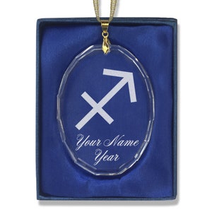 May include: A clear oval crystal ornament with a gold chain and a white Sagittarius zodiac symbol. The ornament is in a blue velvet box with the text "Your Name Year" engraved on the ornament.