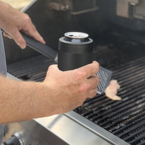 May include: A black insulated can cooler with a silver can inside. The cooler is being held by a person's hand over a grill.