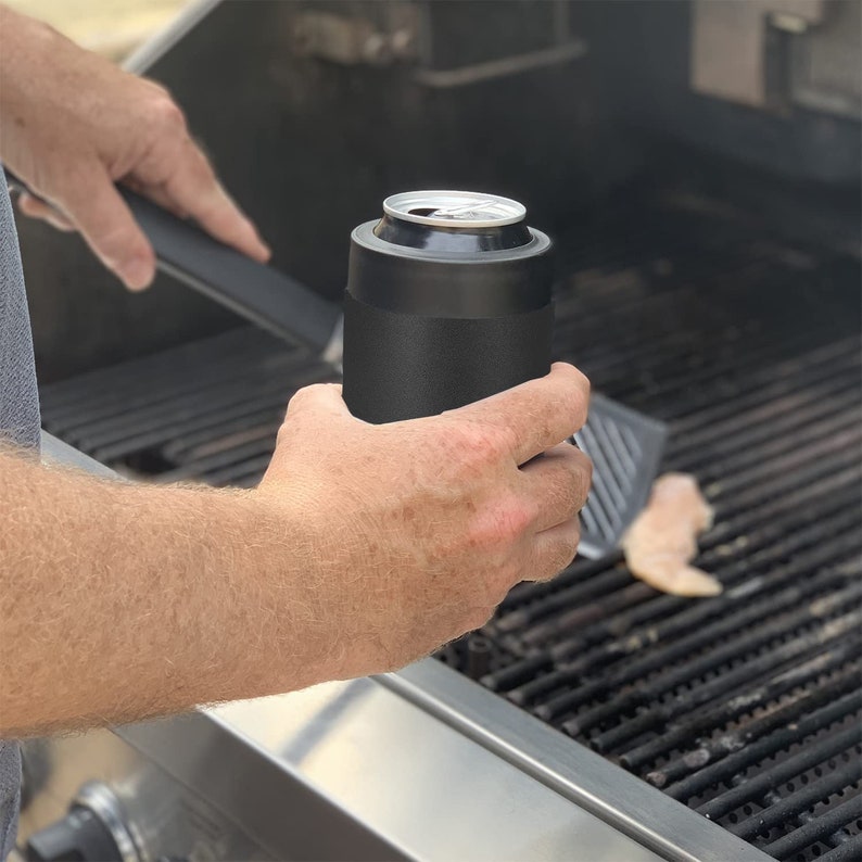 May include: A black insulated can cooler with a silver can inside. The cooler is being held by a person's hand over a grill.