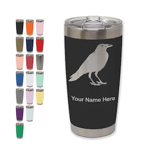May include: A black stainless steel tumbler with a silver raven design and the text "Your Name Here." A variety of colored tumblers are displayed to the left of the main tumbler.