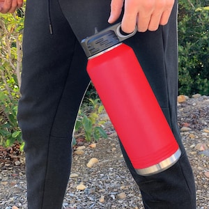 May include: A red stainless steel water bottle with a black lid and a carrying handle. The bottle is being held by a person.