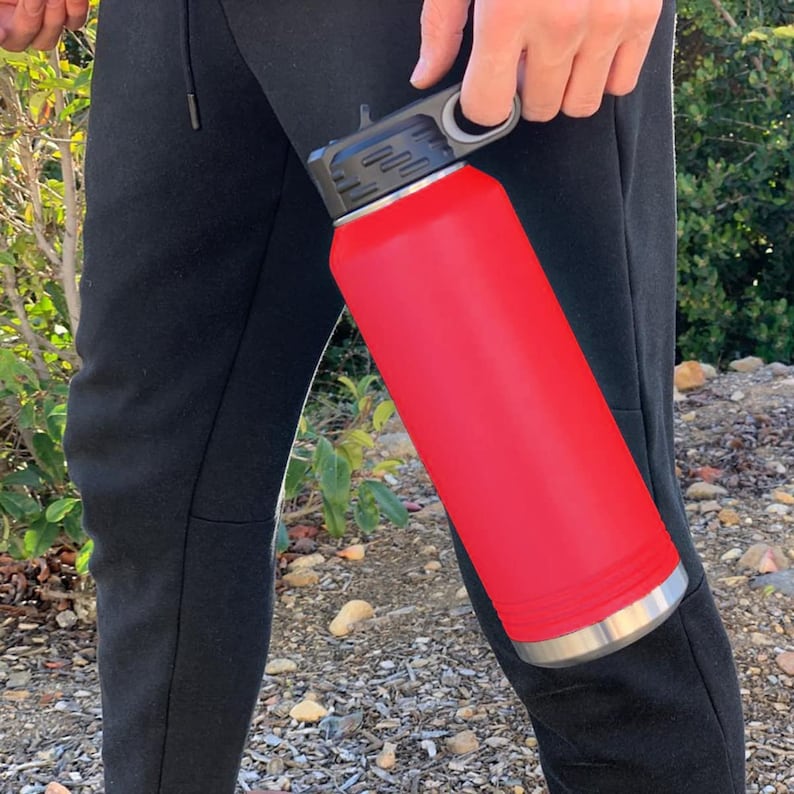 May include: A red stainless steel water bottle with a black lid and a carrying handle. The bottle is being held by a person.