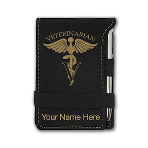 May include: Black leatherette notebook with a gold-colored caduceus symbol and the word "VETERINARIAN" above it. The notebook has a black elastic band closure and a pen holder. The text "Your Name Here" is engraved below the symbol.
