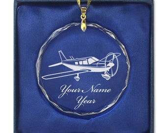 May include: A clear glass bauble with a white silhouette of a small aeroplane and the text "Your Name Year". The bauble is in a blue velvet box.