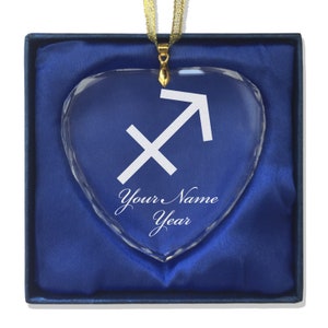May include: A crystal heart ornament with a gold ribbon and a white Sagittarius zodiac symbol. The ornament is in a blue velvet box with the text "Your Name Year" inscribed on the heart.