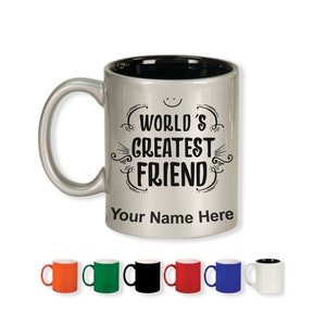 K&ouml;nnte beinhalten: Eine silberne Tasse mit schwarzem Innenraum und gebogenem Henkel. Die Tasse tr&auml;gt den Text "WORLD'S GREATEST FRIEND" in einer dekorativen Schriftart, mit Platz f&uuml;r einen personalisierten Namen. Mehrere farbige Tassen werden darunter angezeigt.