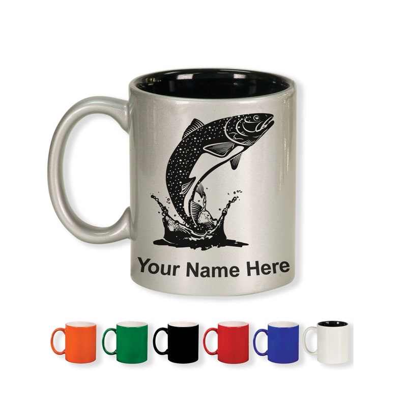 May include: A silver ceramic mug with a black interior and handle. The mug features a black graphic of a fish leaping out of water, with the text "Your Name Here" below. Several colored mugs are displayed below.