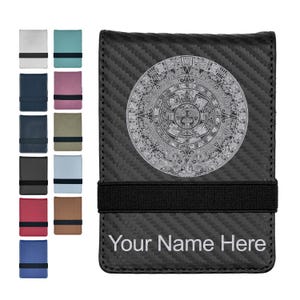 May include: A black carbon fiber wallet featuring a circular Aztec calendar design in white and a horizontal black elastic strap. The text Your Name Here is printed in white at the bottom. A column of ten additional wallet color options is shown.
