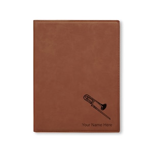May include: A brown leatherette journal with a black silhouette of a trombone and the text "Your Name Here" engraved on the front.