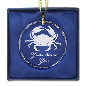May include: A clear glass ornament with a white crab design and the text "Your Name Year" on a blue velvet background.