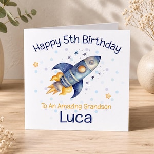 Op de afbeelding: Een vierkante witte wenskaart met een illustratie van een blauwe raket met gele vlammen, omringd door kleine blauwe stippen en sterren. De tekst luidt Happy 5th Birthday, To An Amazing Grandson, en de naam Luca in donkerblauw en geel.