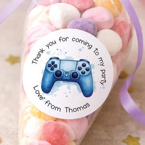 Peut inclure: Un sachet conique en plastique transparent rempli de guimauves aux couleurs pastel, noué avec un ruban violet clair. Une étiquette ronde blanche présente une illustration de manette de jeu bleue et le texte : Thank you for coming to my party. Love from Thomas.