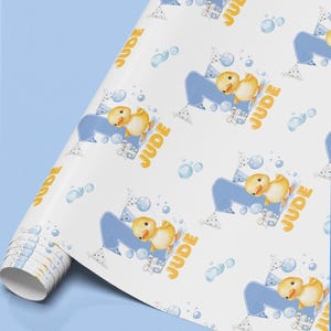 May include: A roll of white wrapping paper featuring a repeating pattern of yellow ducklings, blue number ones, gift boxes, and party bunting. The text JUDE is printed in yellow alongside each duckling, accented by light blue bubbles.