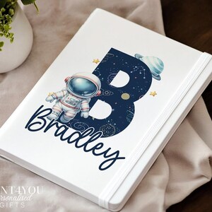 May include: A white notebook featuring a large dark blue letter B filled with space-themed illustrations including stars, planets, and a rocket. An astronaut figure is positioned next to the letter, and the name Bradley is printed in dark blue script.