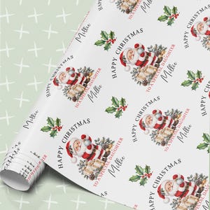 May include: White wrapping paper featuring a repeating pattern of Santa Claus holding a gift, green holly leaves with red berries, and the text Happy Christmas To A Special Daughter Millie in black and red ink on a smooth, matte surface.