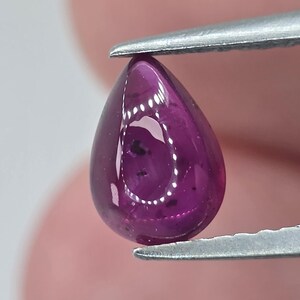 Può includere: Una gemma lucida a forma di goccia, tenuta da pinzette metalliche. La gemma è di un colore magenta intenso e traslucido con sottili inclusioni interne. La superficie riflette la luce, creando riflessi. La gemma è probabilmente un rubino o una pietra preziosa simile.