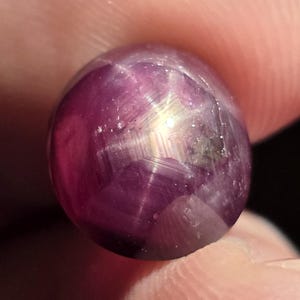 May include: A close-up of a polished, oval-shaped gemstone. The gem is a deep, purplish-red color with a star-like effect visible on its surface. The star pattern is created by light reflecting off of inclusions within the stone.