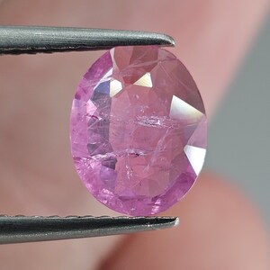 Può includere: Una pietra preziosa ovale di colore rosa con taglio a faccette. La pietra preziosa è tenuta da una pinzetta.
