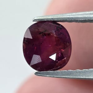 May include: A faceted, oval-shaped, deep red gemstone held by a pair of silver tweezers. The gem has a polished surface with multiple facets that reflect light, creating a sparkling effect. The gemstone's color is a rich, dark red.