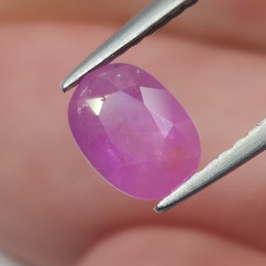 May include: A pink oval-shaped gemstone with a faceted cut. The gemstone is held by tweezers.