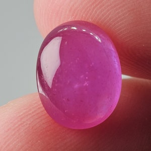 May include: A vibrant, oval-shaped, pink gemstone. The polished surface reflects light, showcasing a glossy finish. The gem is held between fingers, highlighting its size and color. This gemstone could be used in jewelry.