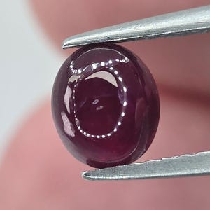 May include: A deep red, oval-shaped gemstone held by a pair of silver tweezers. The polished surface reflects light, creating a bright, circular highlight. The gem's color is a rich, dark shade of red, with a smooth, rounded form.