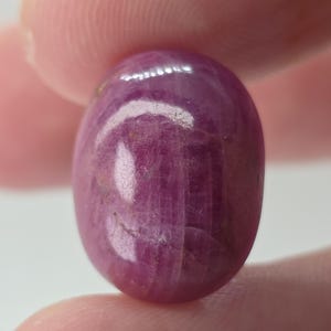 May include: An oval-shaped, cabochon-cut ruby gemstone with a deep magenta-red color. The stone features a smooth, polished surface with visible internal inclusions and natural striations that catch the light, creating a subtle sheen.