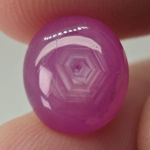 May include: A polished, oval-shaped, pink gemstone with a star-like pattern visible within. The gem is held between fingers, showcasing its vibrant color and internal structure. The gemstone appears to be a ruby.
