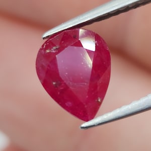 Può includere: Una gemma di rubino sfaccettata a forma di pera, tenuta da una pinzetta. Il rubino è di colore rosso intenso con inclusioni visibili. La gemma è tagliata per massimizzare la riflessione della luce, mostrando la sua brillantezza.