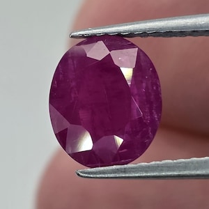 May include: A faceted, oval-shaped ruby gemstone held by tweezers. The ruby is a deep, rich red color with multiple facets that reflect light. The gemstone is a focal point, showcasing its clarity and brilliance.