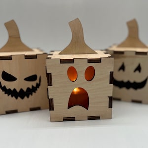 May include: Three wooden jack-o'-lantern boxes with different facial expressions. The box in the center has a lit candle inside, creating a warm glow.