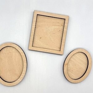 May include: Three unfinished wooden frames in different shapes: an oval, a square, and another oval. The frames have a natural wood color with a dark border. The objects are arranged on a white surface.