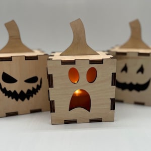 Pode incluir: Três caixas de madeira de Halloween em forma de abóbora com diferentes expressões faciais. As caixas são feitas de madeira clara e têm um design recortado. A caixa da esquerda tem uma cara assustadora, a caixa do meio tem uma cara de surpresa e a caixa da direita tem uma cara feliz.