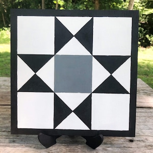 May include: Black and white quilt block with a gray center square. The block is made of squares and triangles and is displayed on a wooden stand.