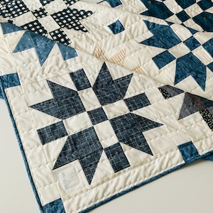 May include: A handmade quilt featuring a starburst pattern in shades of blue and white. The quilt has a patchwork design with various fabric textures, including gingham and floral prints. The edges are finished with a blue floral border and white binding.