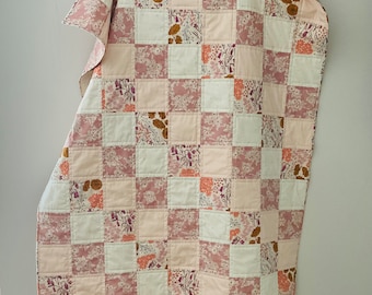 Könnte beinhalten: Eine rosafarbene und weiße Patchwork-Decke mit Blumenmuster. Die Decke besteht aus Quadraten aus verschiedenen Stoffen, die jeweils ein anderes Blumenmuster aufweisen. Die Decke ist gefaltet und auf einem Holzständer ausgestellt.